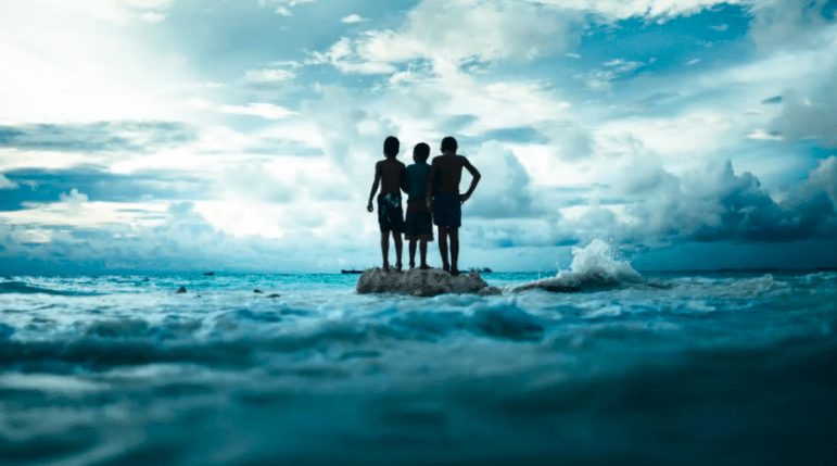 Three people stand on a rock surrounded by small waves sillouetted against a cloudy sky, the whole scene is quite blue.