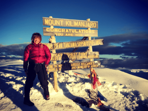 A person in a red jacket stands surrounded by snow next to sign reading "Mount Kilimanjaro" with many additional signs below.