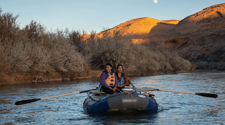 Two people float in a dark inflatable raft with oars extended, brush lines the shore and fading light catches the hills in the background.