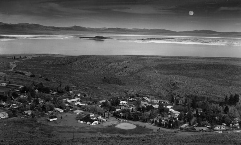 A black and white photo of the small town of Lee Vining, taken from a hill above, showing about 30 homes and shops with Mono Lake and black Negit Island and white Paoha Island, and the full moon rising in the distance.